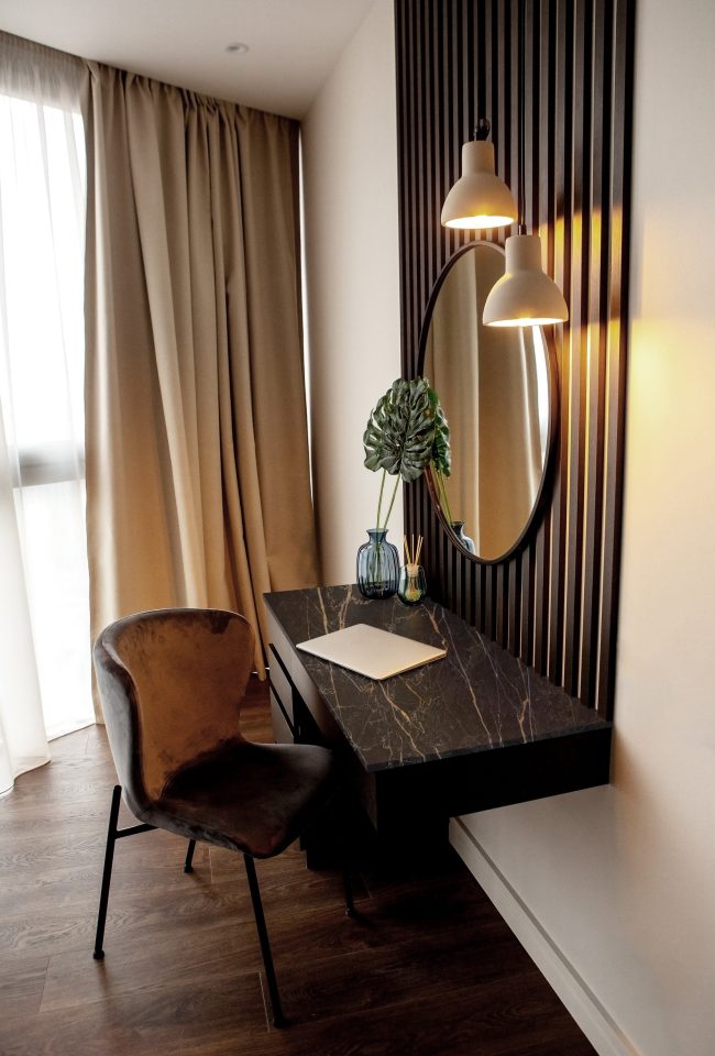 Dark interior. Cozy workplace in the apartment in the bedroom near the window. Stylish round mirror in a wooden frame above the table. On the table is a laptop, a vase with a flower and a fragrance